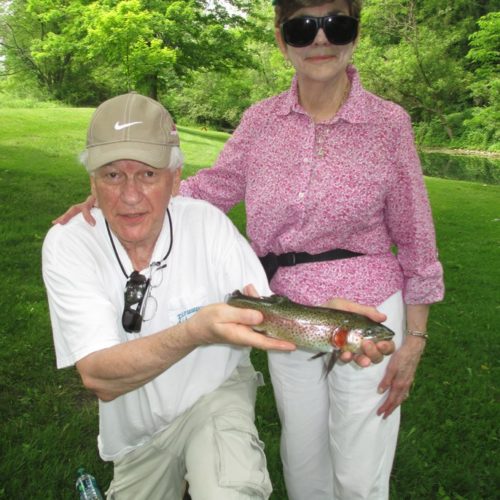 An older man kneeled down holding his trout next to his wife standing beside him