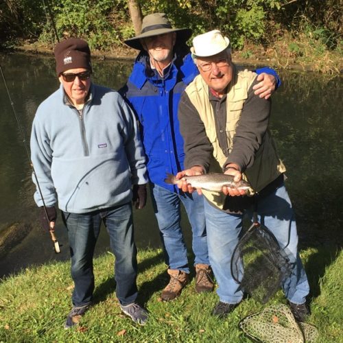 Three older men standing together holding a trout by the creek bank