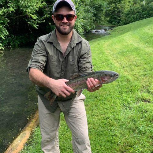 A young man holding a rainbow trout standing on the creek bank