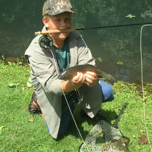 A young boy holding the rod in his mouth while kneeled down and posing for a photo with the trout he caught