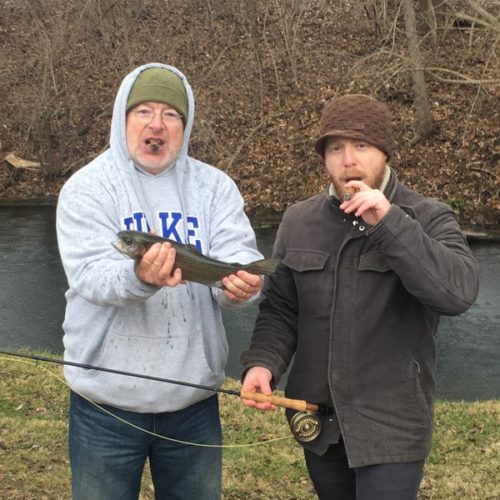 An older gentleman and middle-aged man smoking cigars while posing for a photo with a trout