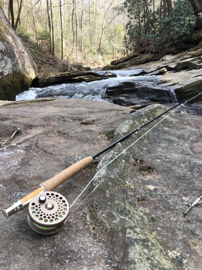 A close-up photo of a fly fishing rod and reel sitting on a rock in the middle of a stream