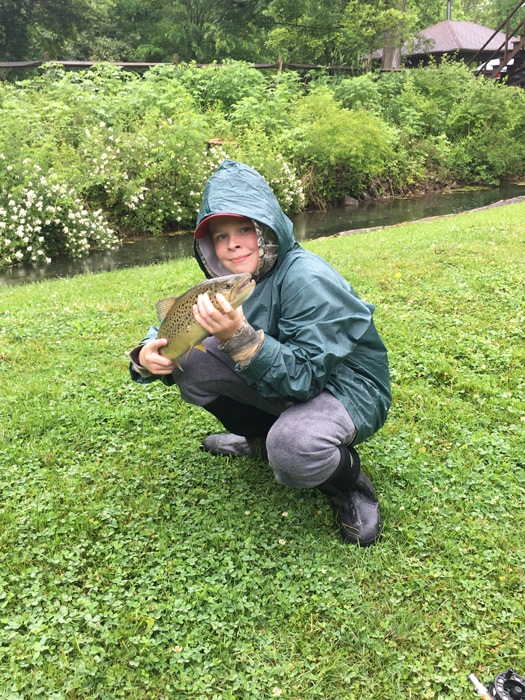 A young boy in a rain coat kneeled down on the bank holding a brown trout.