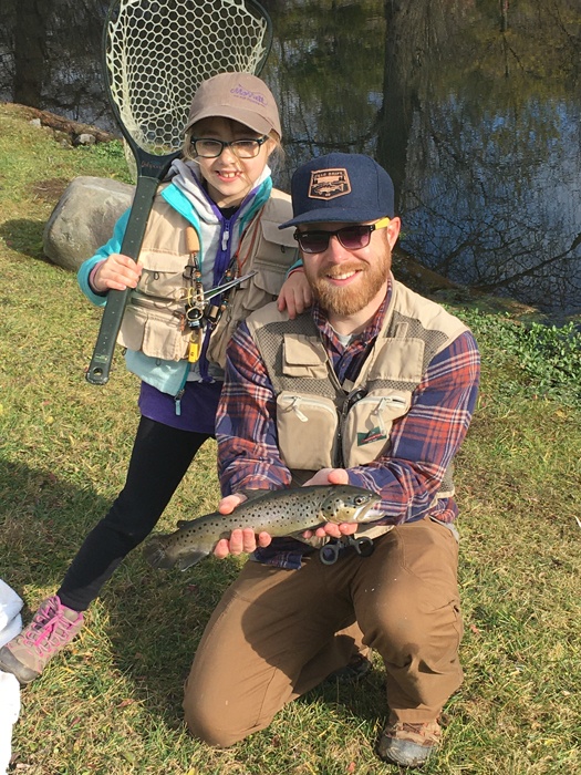 Eva standing with net in hand behind Andy crouched down holding a trout.