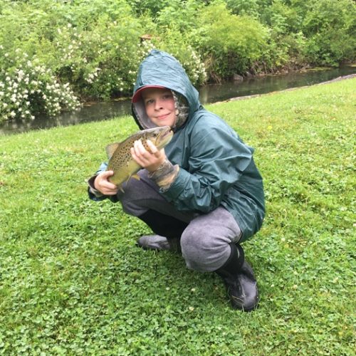 A young boy crouched down in a raincoat posing with his trout.