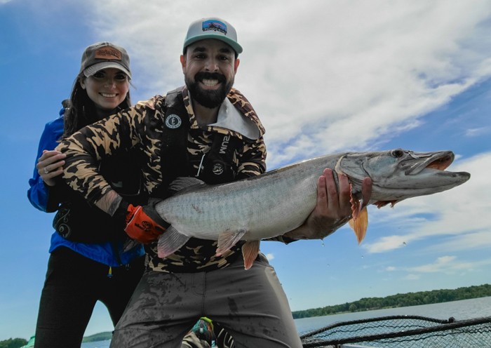 Nicole and Ben standing in their fishing boat holding a muskie.