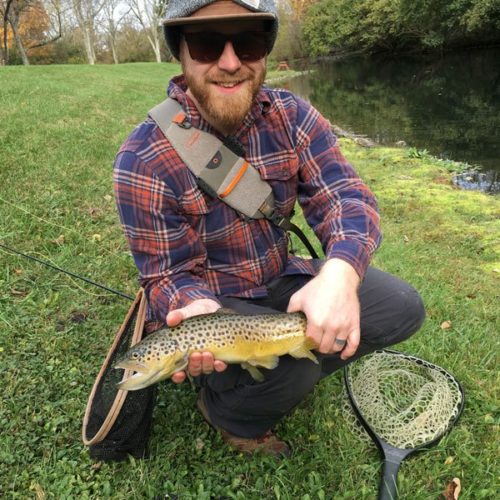 A young man kneeled down holding a brown trout wearing sunglasses and a beanie hat