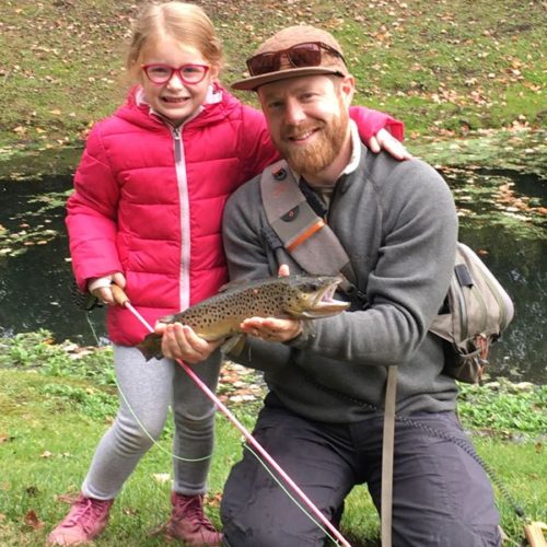 A young girl standing next to her daddy kneeled down holding a brown trout