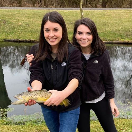 Two young ladies standing while one of them holds a brown trout